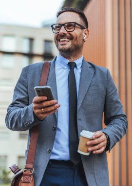 Elinde kahve fincanı ve akıllı telefon tutarken gülümseyen takım elbiseli profesyonel bir adam, kulaklıklarla dışarıda yürüyor. Kentsel bir ortamda başarılı iş, güven ve modern yaşam tarzını temsil eder.