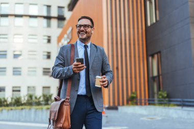 Takım elbiseli neşeli bir işadamı elinde kahve fincanı ve akıllı telefonuyla modern bir ofis binasının önünde yürüyor. Kendine güveni ve başarısı var, işte verimli bir günün tadını çıkarıyor..