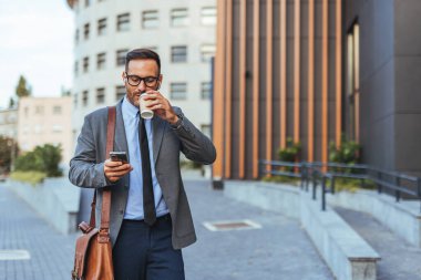 Kendine güvenen bir işadamı kahve içerken ve telefonunu kontrol ederken şehir merkezinde yürüyor. Deri bir evrak çantası taşıyor, modern, profesyonel şehir hayatının vücut bulmuş hali..