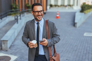 Kendine güveni tam bir işadamı elinde kahve ve akıllı telefonla dışarı çıkıyor, neşeli bir şekilde gülümsüyor. Takım elbise ve kravat takmış, modern şehir ortamında yakalanmış, profesyonel ve olumlu bir ruh hali içinde..