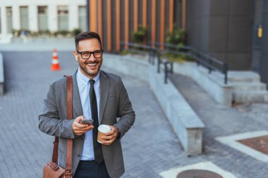 Takım elbiseli gülümseyen iş adamı elinde kahve ve akıllı telefonuyla dışarıda dolaşıyor. Gözlük takıyor ve kendinden emin görünüyor, başarıyı ve mutluluğu temsil ediyor..