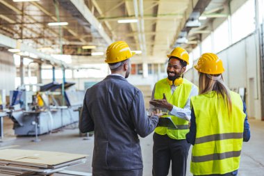 Çağdaş bir fabrika ortamında bir projeyi tartışan güvenlik donanımlı çeşitli profesyonellerden oluşan bir grup, takım çalışması, yenilik ve endüstriyel işbirliği sergiliyor..