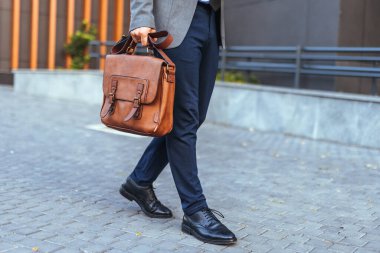 Resmi giyinmiş bir işadamı, elinde şık bir deri evrak çantası olan kaldırımlı bir cadde boyunca güvenle yürüyor, profesyonellik ve kentsel yaşam tarzını sergiliyor. Ayarlar güven ve modernlik yayar..