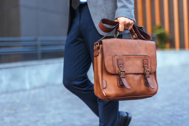 İyi giyimli bir profesyonel şık bir kahverengi deri evrak çantası taşıyor, şehir merkezindeki bir iş ya da kurumsal çevreyi tasvir ediyor. Profesyonellik ve moda temaları için ideal.