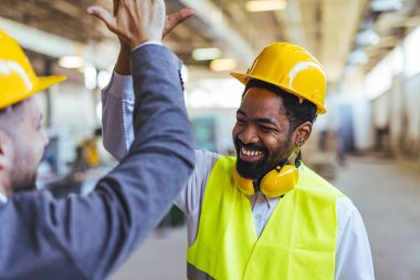 Güvenlik kaskı ve ekipmanı takan iki inşaat işçisi, iyi aydınlatılmış bir endüstriyel atölyede gülümseyip beşlik çakıyor, takım çalışması, başarı, işbirliği ve olumlu atmosfer yayıyor, başarı ve profesyonel arkadaşlığı resmediyorlar..