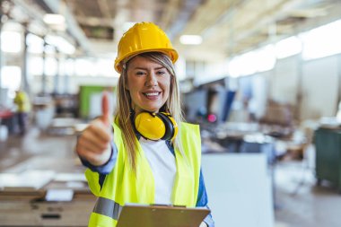 Kendine güvenen kadın inşaat işçisi güvenlik kıyafeti giyiyor, gülümsüyor ve endüstriyel ortamda onay veriyor. Profosyonelliği, güvenliği ve inşaat ve sanayide olumlu çalışma ortamını teşvik eder.