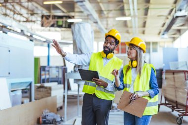 Fabrika ortamındaki planları gözden geçiren iki yüksek görüşlü yelek ve başlık mühendisi, makine ortamında takım çalışması ve endüstriyel işbirliği gösteriyorlar..