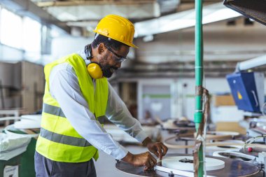 Koruyucu ekipman giyen erkek teknisyen, kalite ve güvenliği vurgulayarak endüstriyel bir ortamda bir makinede çalışıyor. Üretim süreçlerini ve üretim uzmanlığını profesyonel bir ortamda gösterir.