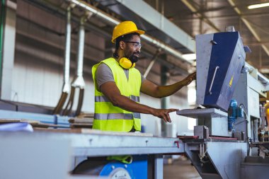 Büyük bir imalat tesisinde çalışan sanayi işçisi, iş güvenliği ve uygun düzenlemelere uymak için sıkı bir şapka, eldiven ve yüksek görünürlüklü yelek giyerken aynı zamanda uygun güvenlik teçhizatı giyer..