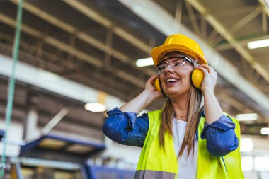 Bir fabrika ortamında, koruyucu gözlük takan ve kulaklık takan bir bayan işçi gülümsüyor. Endüstriyel bir işyerinde güvenlik, pozitiflik ve profesyonellik göstergesi.