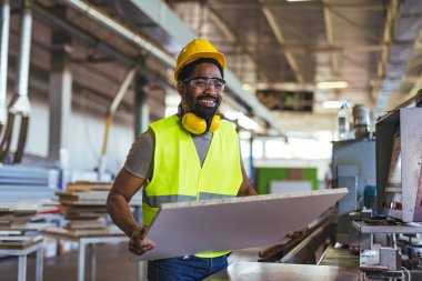 Yüksek görüş açısına sahip yelek ve güvenlik teçhizatıyla çalışan işçiler bir fabrikada inşaat panelini güvenli bir şekilde manevra ettiriyor..