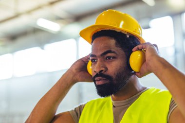 Kulak koruyucu takan ve kasketli erkek inşaat işçisi fabrika ortamında güvenliği sağlıyor. İmaj, mesleki güvenliğe odaklanarak profesyonellik, odaklanma ve işyeri sorumluluğunu aktarmaktadır..
