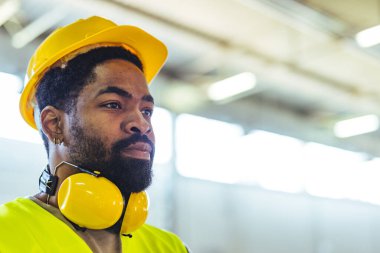 İnşaat işçisi, sanayi ortamında güvenlik ve profesyonelliği sembolize ederek, kararlılığı vurgulayarak ve işyeri sağlık ve güvenlik uygulamalarına odaklanarak, sıkı bir şapka ve kulak koruyucusu takıyor..