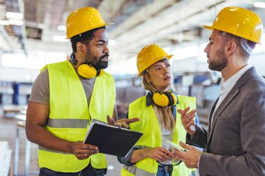 Güvenlik teçhizatı taşıyan üç inşaat işçisi, sanayi işyerleri için gerekli takım çalışması, işbirliği ve güvenlik protokollerine odaklanarak geniş bir deponun içinde bir görüşme yaptılar..