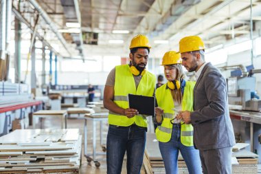 Endüstriyel bir ortamda çalışan çeşitli profesyonellerden oluşan bir grup, güvenlik teçhizatı ve aletlerle donatılmışken süreç ayrıntılarını tartışıyor. Modern bir üretim ortamında işbirliği ve takım çalışması.