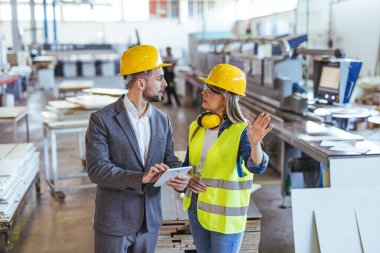 Güvenlik ekipmanı kullanan iki profesyonel, bir sanayi fabrikasında takım çalışması ve lojistik ürünleri imalatında işbirliği yapıyor..