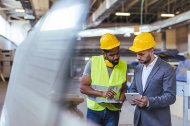Üretim ortamında güvenlik teçhizatı projesi planları giyen iki işçi. Görüntü, endüstriyel bir ortamda işbirliği ve işyeri iletişimini yakalıyor.