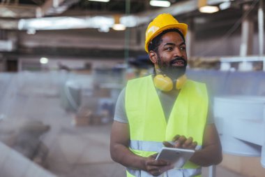 Bir kask ve yelek de dahil olmak üzere güvenlik teçhizatı kullanan bir sanayi işçisi, endüstriyel süreçler için yenilik, güvenlik ve dijital araçları vurgulayan bir imalat ortamında modern teknolojiyle çalışır..