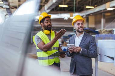 Güvenlik kaskı ve yelek giyen iki profesyonel, dijital tablet ve not panosuyla bir fabrikada işbirliği yaparak bina planlarına odaklanıyor. İnşaat ve imalatta takım çalışmasını, üretkenliği ve yeniliği sembolize ederler..