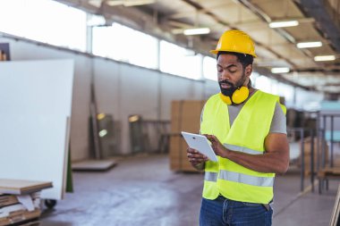 Güvenlik yeleği ve kask giyen adam büyük endüstriyel tesislerde tablet kullanıyor. Konseptler arasında teknoloji, modern imalat ve işyeri güvenliği yer alıyor.