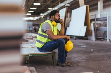 Yansıtıcı yelek ve kulaklık takan bir işçi atölyede elinde kaskla dalgın dalgın oturur. Arka plan sanayi ortamını gözler önüne seriyor, işçilik, güvenlik ve adanmışlık vurgulanıyor.