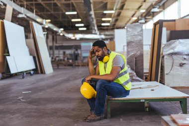 Fabrika işçisi atölyedeki bir panelde dinleniyor. Yorgunluğunu dile getiriyor ve elinde şapka tutuyor..