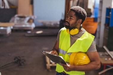 Güvenlik yeleği giymiş bir işçi elinde bir pano ve miğfer tutarak bir depoda durup çevreyi dikkatle izliyor. Görüntü, inşaat ortamlarında sanayi, odak ve güvenlik protokollerini vurguluyor.