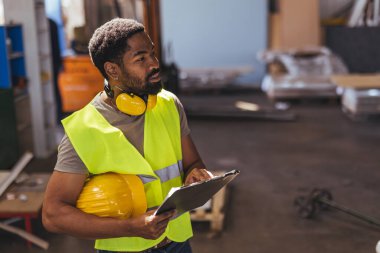 Endüstriyel çalışma alanlarında güvenlik ve organizasyonu simgeleyen bir atölyede koruyucu ekipman ve pano ile odaklanmış inşaat işçisi.