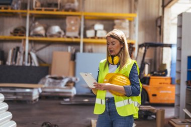 Güvenlik teçhizatı olan bir depo işçisi rafların ve aletlerin arasında envanter yönetimi için tablet kullanır. Teknolojinin modern endüstriyel ortamlardaki rolünü göstermek, verimliliği ve yeniliği vurgulamak.