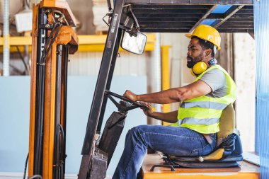 Bir depo işçisi sarı bir başlık, yansıtıcı güvenlik yeleği ve teçhizat giyerken forklift kullanır. Sanayi ortamında, yoğun bir lojistik ortamda profesyonellik ve işyeri güvenliği vurgulanıyor.