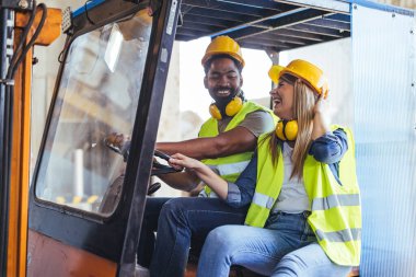 Canlı güvenlik kıyafeti giyen iki inşaat işçisi sarı bir forklift kullanarak mutlu bir anı paylaşıyorlar..