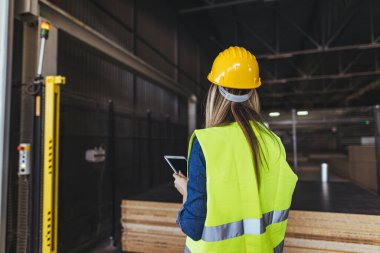 Yüksek görüş açısına sahip yelek ve başlıklı profesyonel kadın danışman, organize depolama tesislerindeki iş akışlarını yönetmek ve denetlemek için dijital tablet kullanarak endüstriyel depo operasyonlarını inceliyor..