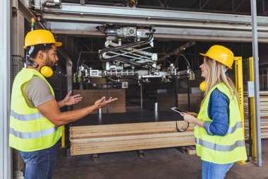 Güvenlik teçhizatı taşıyan iki işçi bir marangozluk tesisinde makine operasyonlarını tartışıyor..