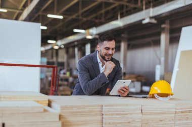Bir üretim tesisinde profesyonel mimar, bir tablet üzerindeki tasarımları incelerken proje üretim süreçlerini denetliyor, liderlik, yenilik ve endüstriyel mühendislik gelişmelerini tasvir ediyor..