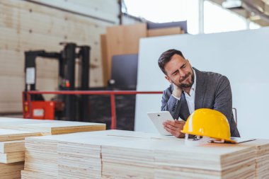 Profesyonel bir mimar proje planlarını inşaat ortamında tablet kullanarak inceler ve gözle görülür bir boyun ağrısı yaşar. Sert şapka ve endüstriyel arka plan inşaat ve mühendisliği sembolize ediyor..