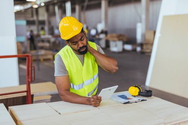 Kask ve güvenlik yeleği giyen bir fabrika işçisi, bir tablet üzerinde boynunu esnerken planlarını gözden geçirir. Bu da profesyonel bir imalat ortamında gerginlik ve çabayı gösterir..