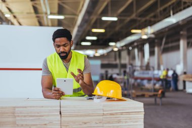 Endüstriyel bir ortamda çelik yelek giyen bir erkek işçi görev için tablet kullanır. Yakınlarda sarı bir başlık var. Üretim yerlerinde güvenlik standartlarına uyulduğunu vurguluyor..