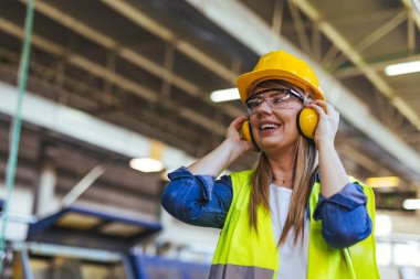 Neşeli bir kadın inşaat işçisi olay yerinde kask ve kulak koruyucusu da dahil olmak üzere koruyucu giysi giyer. Fotoğraf, iş yerinde güvenlik ve profesyonelliğin özünü yansıtıyor..