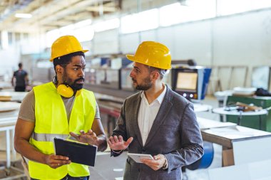 Güvenlik donanımı giyen iki profesyonel endüstriyel üretim binasında bir projeyi tartışıyor..