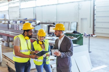 Mühendisler ve bir işletme profesyoneli, takım çalışması ve üretkenliği vurgulayarak modern bir endüstriyel tesiste işbirliği yapıyorlar. Güvenlik ekipmanları ve belgeler, üretim ve planlamaya yönelik organize bir yaklaşımı vurguluyor.