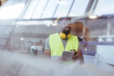 Koruyucu giysiler giyen ve elinde tablet tutan bir fabrika işçisi iyi aydınlatılmış bir endüstriyel ortamda gülümsüyor, verimlilik ve pozitif çalışma ortamı gösteriyor..