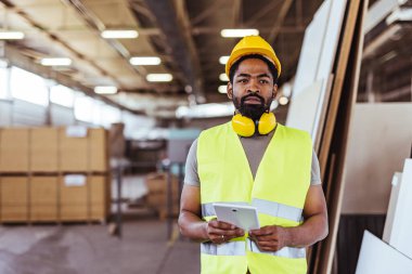Sert şapka ve yansıtıcı yelek giyen bir işçi, sanayi ortamlarında profesyonelliği, güvenliği ve modern teknolojiyi simgeleyen endüstriyel ekipman ve malzemelerle çevrili geniş bir kapalı depo deposunda bir tablet tutuyor..