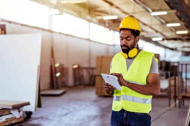 Yansıtıcı teçhizat ve kask takan bir işçi, büyük ekipman ve malzemelerle dolu geniş bir sanayi deposundaki görevleri yöneten bir tablet cihazıyla etkileşim halindedir. Çevre verimliliği ve organizasyonu simgeler.