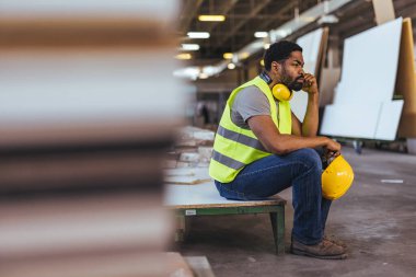 Yansıtıcı yelek giyen bir inşaat işçisi bir depoda oturuyor, bir kask tutuyor ve düşünceli görünüyor, sıkı çalışma, tefekkür ve güvenlik kavramlarını tasvir ediyor..