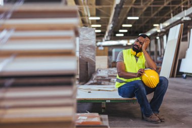Çalışan bir imalat atölyesinde güvenlik yeleği giyiyor, dinlenirken yorgunluk ve gerginlik belirtileri gösteriyor, iş stresi ve önlemleri vurguluyor.