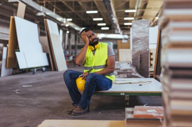 Fabrikada oturan yorgun bir inşaat işçisi, endüstri malzemeleri ve araçlarıyla çevrili, endüstriyel ortamlarda hayal kırıklığını, aşırı çalışmayı ve işyeri streslerini temsil eden bitkinlik içinde başını sıkıca tutuyor..