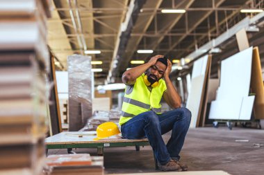 Yansıtıcı yelek giyen ve fabrikanın zemininde oturan bir işçi. Stresli ve bunalmış görünüyor. Yanında koruyucu bir şapka ve kulaklıkla..