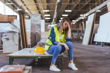 Molada kadın fabrika işçisi, stres ve yorgunluk gösteriyor, güvenlik ekipmanları ve aletlerinin yanında oturuyor, endüstriyel ortamda. Üretimde işyeri sorunları ve güvenlik önceliklerini yansıtır.