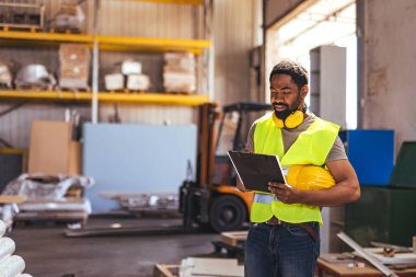 Depo işçisi yüksek görüş açısına sahip yelek giyiyor ve yoğun bir endüstriyel ortamda belgeleri incelerken koruyucu ekipman tutuyor. Arkaplanda organize bir çalışma alanındaki raf, ekipman ve çalışma unsurları gösteriliyor.