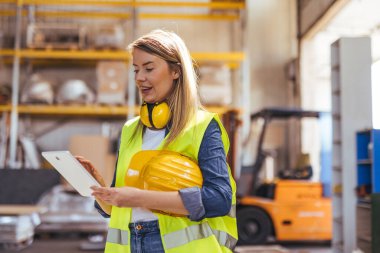 Koruyucu teçhizat tutan ve endüstriyel ambardaki tablet üzerindeki verileri kontrol eden kendine güvenen bir kadın işçi. Parlak sarı başlık ve yelek endüstriyel depolama ortamında güvenlik uygulamalarını vurguluyor.